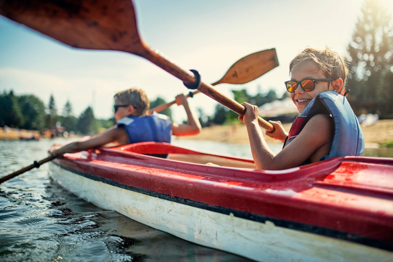 Canoewintercamp Two children sitting in a canoe on water, holding oars and wearing sunglasses and life jackets.